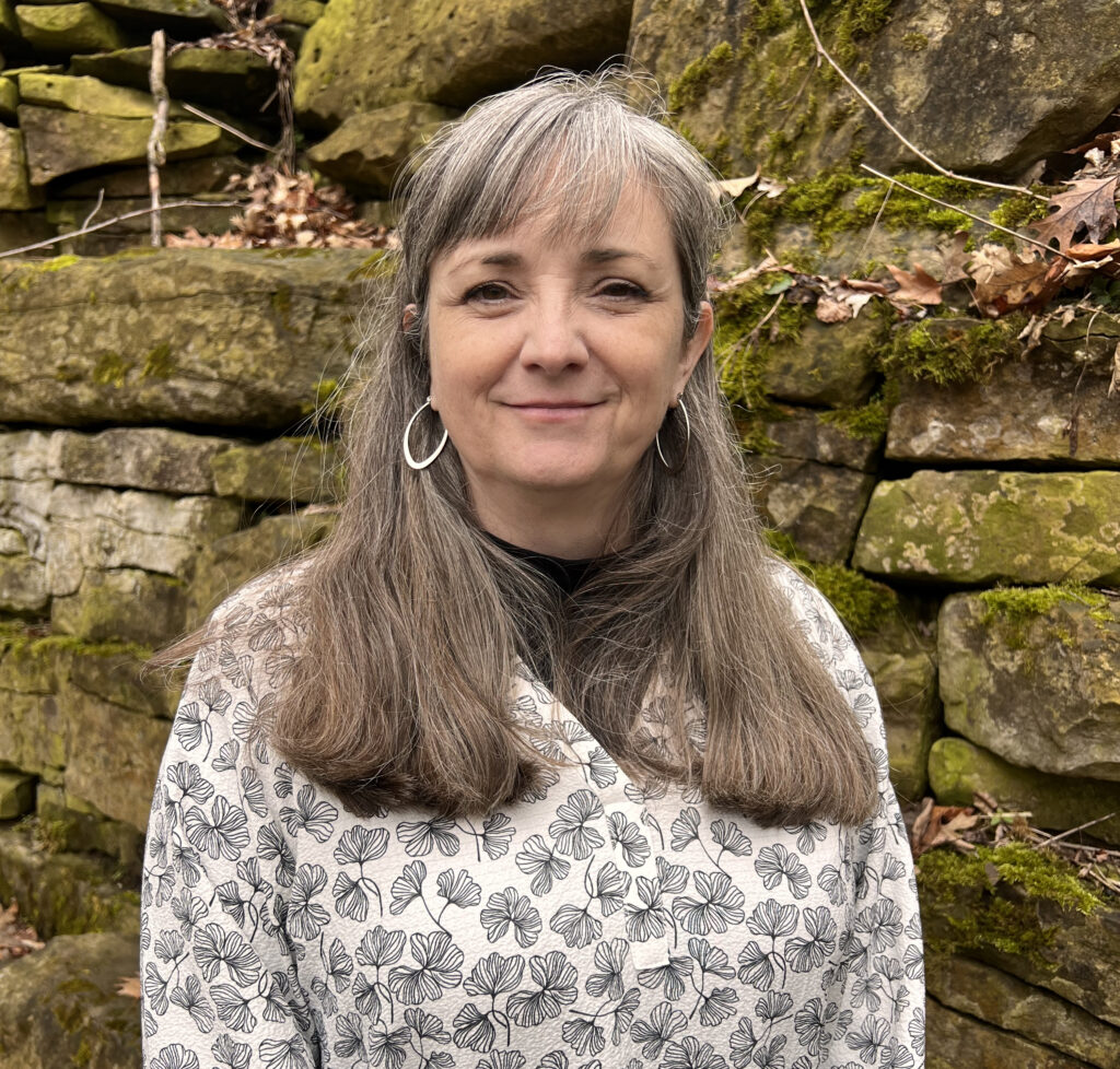 Alisa Cunnington in front of a mossy rock wall wearing a black and white leaf-printed long-sleeved top with a black shirt underneath, silver hoop earrings, and smiling into the camera.]