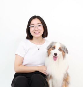 Jenny Ge with her dog River. Jenny is wearing black pants, a white top, and silver horn-rimmed glasses, and River has white fur with light-brown and grey patches and his tongue sticking out