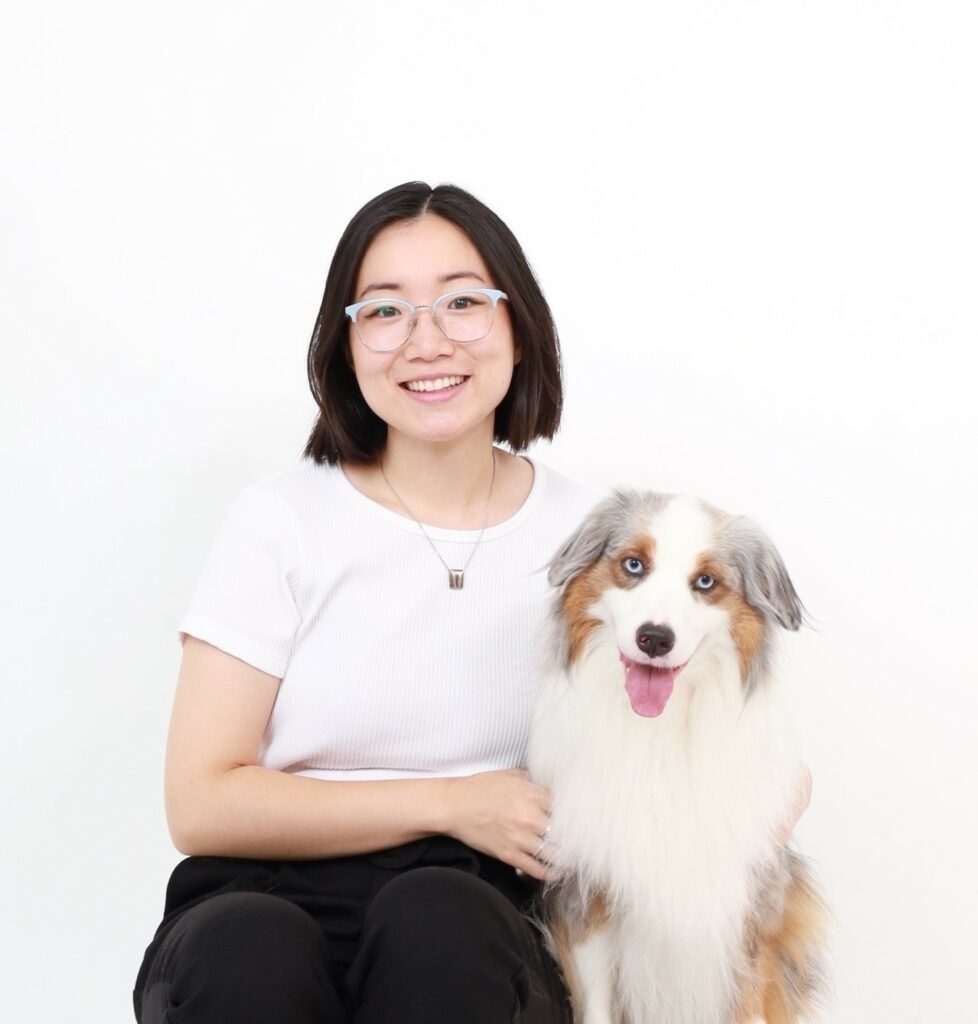 Jenny Ge with her dog River. Jenny is wearing black pants, a white top, and silver horn-rimmed glasses, and River has white fur with light-brown and grey patches and his tongue sticking out