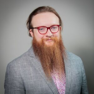 Matthew Dunleavy wearing a pink and purple polka-dot shirt under a grey blazer with red-framed glasses and a long reddish-brown beard smiling into the camera