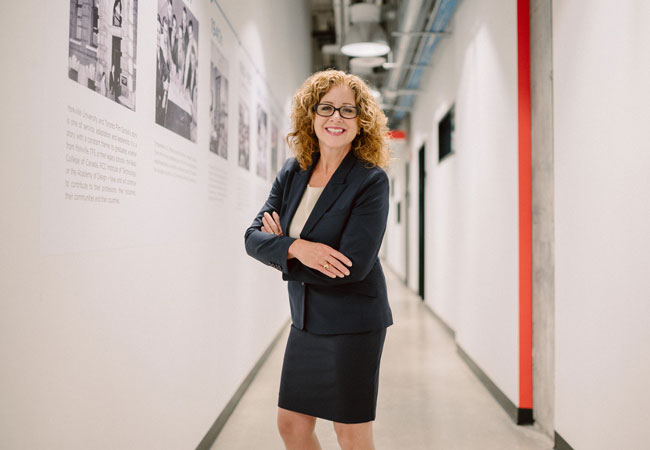 Julia wearing navy blue and standing with her arms folded across her chest, smiling into the camera, and standing in the TFS Alumni Hall at Toronto Film School.